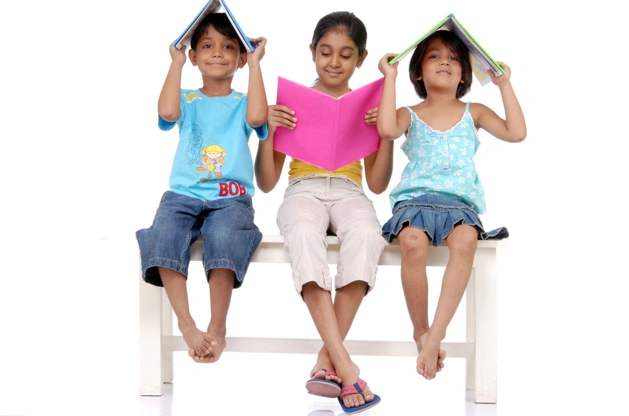 Three children sitting on a bench with books over their heads.